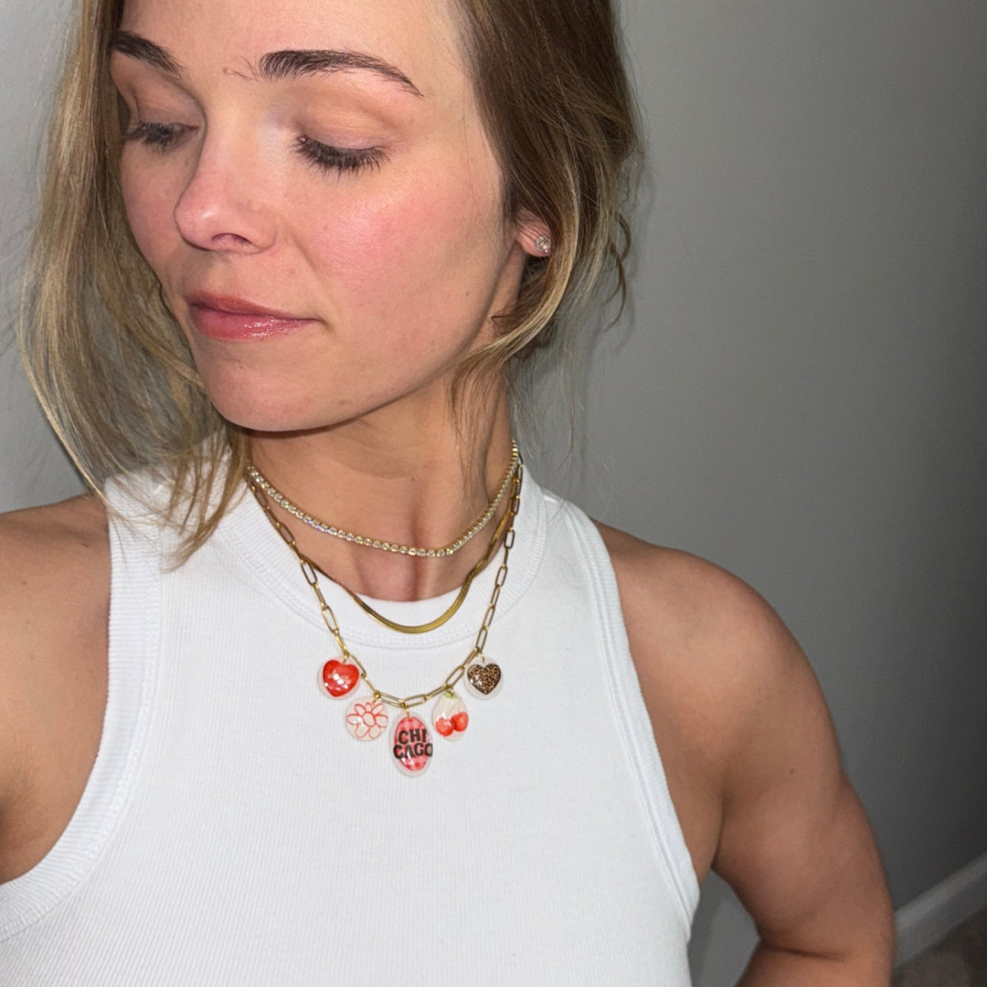 Woman wearing a white tank top and multiple necklaces with heart pendants against a plain background.