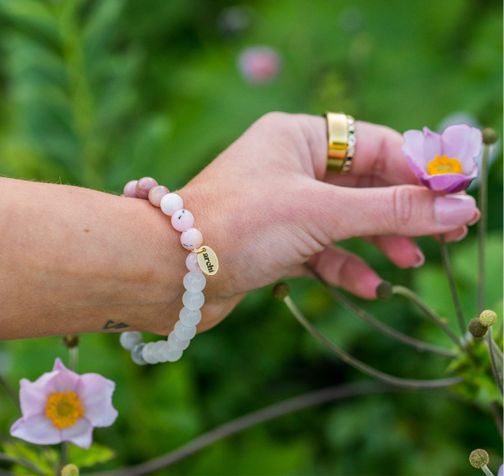 Amara Self Love Bracelet with light pink, sage green, pearl white, and soft tan beads on a wrist surrounded by flowers.